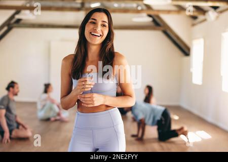 Eine Yoga-Lehrerin lächelt, während sie in einem Yoga-Studio steht, mit ihrer Klasse im Hintergrund. Glückliche junge Frau, die eine Trainingseinheit mit einem G hat Stockfoto