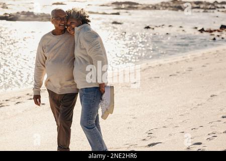 Glückliches älteres Paar, das lächelt und sich umarmt, während man barfuß am Strand spaziert. Ein romantisches Seniorenpaar, das eine schöne Zeit miteinander verbringt Stockfoto