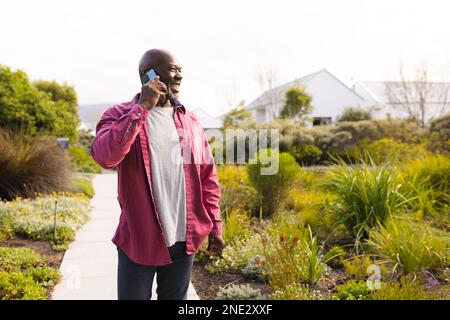 Ein afroamerikanischer Seniorist lächelt, während er auf dem Gartenweg mit dem Smartphone spricht. Altersruhestandsleben und Lebenskonzept Stockfoto