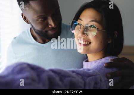 Glückliches, vielseitiges Paar, das umarmt und durch das Fenster schaut. Verbringen Sie Zeit zu Hause, in Ihrem Haushalt und in Ihrem Lebensstil. Stockfoto