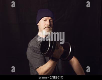 Fitness, ein älterer Mann mit grauem Bart trainiert Bizeps mit Kurzhanteln. Stockfoto