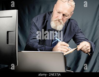Im Büro schreibt ein ernster älterer Mann mit grauem Bart vor einem Laptop in ein Notizbuch. Stockfoto