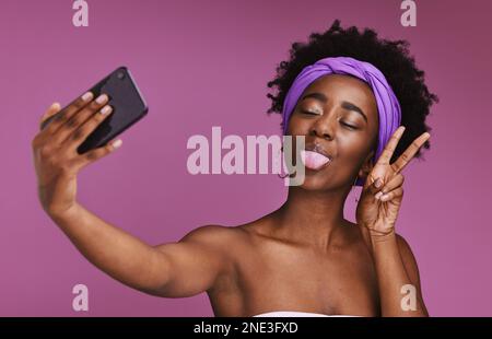 Telefon, Selfie und Gesicht einer schwarzen Frau mit Friedenszeichen für Wellness, Mode und Kosmetik auf lila Hintergrund. Schönheit, Hautpflege und Beeinflussung von Mädchen Stockfoto