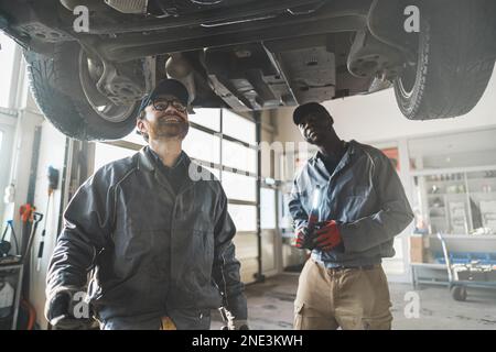 Mittellange Aufnahme von zwei Mechanikern, die die Unterseite des Autos inspizieren. Hochwertiges Foto Stockfoto