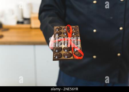 Die Hand des männlichen Konditors hält einen dunklen Schokoriegel mit rotem Band. Hochwertiges Foto Stockfoto