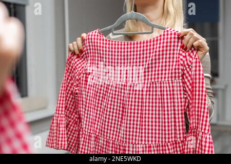 Eine Frau nimmt ein rotes Kleid aus dem Schrank. Eine Frau, die ein Sommerkleid anprobiert. Eine Frau, die vor einem Spiegel steht Stockfoto