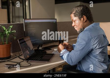 Schwarzer Mann schreibt Code-Zeilen auf einem Desktop-PC mit zwei Monitoren und einem Laptop in einem stilvollen Büro. Professionelle Programmierung Männlicher Entwickler Stockfoto