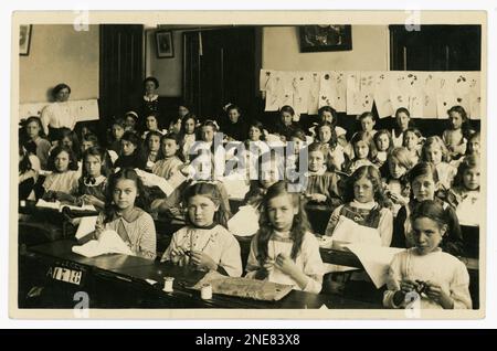 Originelle und klare Postkarte aus dem WW1. Jahrhundert mit Mädchen aus der Juniorschule in einem Nähkurs, mit Samplern, die möglicherweise Stickerei praktizieren, einige stricken, viele Charaktere. Keine Uniformen. Naturzeichnungen an der Wand. Zwei Lehrerinnen schauen sich das an. Aus dem Studio von J.& G Taylor, Green Lane, N. London, Großbritannien Etwa 1913-1919. Stockfoto