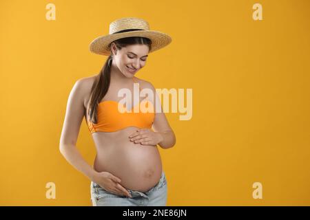 Junge schwangere Frau, die Badeanzug auf gelbem Hintergrund trägt, Nahaufnahme. Sonnenschutzkonzept Stockfoto