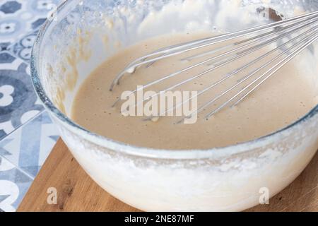 Teig in einer Glasschüssel mit einem Schneebesen aus Metall mischen. Auf einem Holzbrett eine blau geflieste Arbeitsplatte Stockfoto