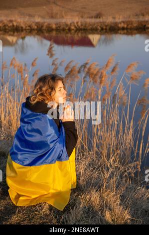 Eine traurige junge Patriotin, umhüllt von einer ukrainischen Flagge, sitzt verzweifelt am Ufer eines Sees und betet für Frieden in der Ukraine. Russische Aggression. - ja, ich weiß Stockfoto