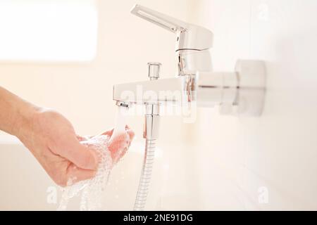 Männliche Hand prüft die Temperatur des fließenden Wassers aus einem Wasserhahn in einem hellen Badezimmer- selektiver Fokus Stockfoto