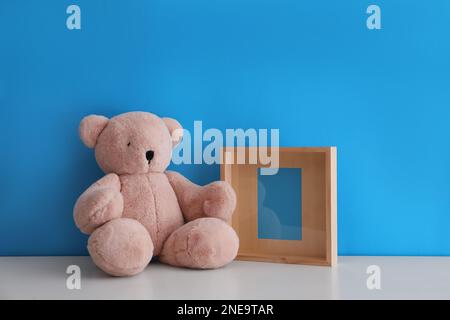 Leerer Bilderrahmen und Teddybär auf weißem Tisch in der Nähe der hellblauen Wand. Raum für Design Stockfoto