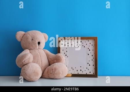 Leerer Bilderrahmen und Teddybär auf weißem Tisch in der Nähe der hellblauen Wand. Raum für Design Stockfoto