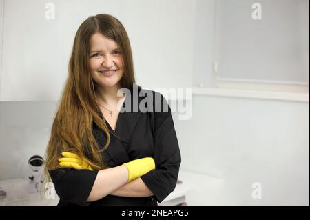 Wunderschöne junge Frau in gelben Latexhandschuhen mit fließendem Haar und in einem schwarzen Bademantel auf weißem Hintergrund Hände des Küchenchefs. Küchenhygiene. Frau trägt schwarze Latexhandschuhe, isoliert mit Licht Stockfoto