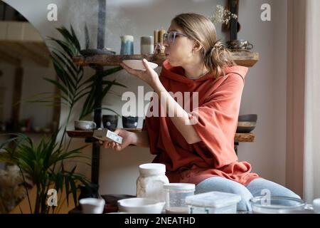 Die meisterhafte Töpferin bläst Staub ab, nachdem sie handgefertigte Tonprodukte gereinigt hat. Vom Hobby bis zum Kleinunternehmen Stockfoto