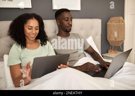 Lächelndes multirassisches Paar, das morgens im Bett verschiedene Geräte benutzt Stockfoto