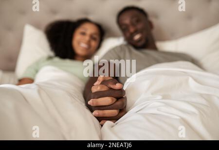 Lächelndes, junges, multiethnisches Paar, das sich im Bett die Hände hält Stockfoto