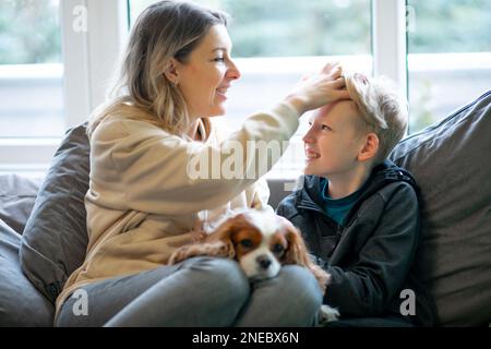 Porträt einer glücklichen, schönen Frau mittleren Alters, die mit einem Teenager zu Hause auf dem Sofa sitzt, den Hund hält, den Kopf des Sohnes streichelt. Stockfoto