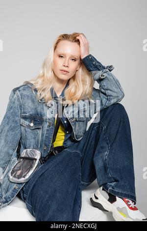 Blonde Frau in blauem Denim-Outfit und trendigen Jeans auf weißem Würfel isoliert auf grauem Stockbild Stockfoto