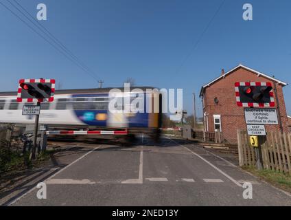 24. März 2022 - Ein fahrender Zug, der an klaren Tagen mit blauem Himmel über eine automatische Halbbarriere fährt. Stockfoto