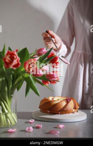 Eine Frau, die einen Kuchen mit Glasur dekoriert. Strauß Tulpenblumen und gebündelter Kuchen auf dem Tisch Stockfoto