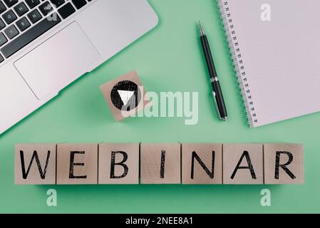Word WEBINAR und Play Button Symbol auf Holzwürfeln vor grünem Hintergrund mit Laptop und Notizblock, E-Learning und Bildungskonzept Stockfoto