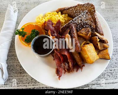 Nahaufnahme eines großen Frühstückstellers mit Rührei, Speck, französischem Toast, Würstchen, Kartoffeln, Ahornsirup und eine Orangenscheibe. Stockfoto