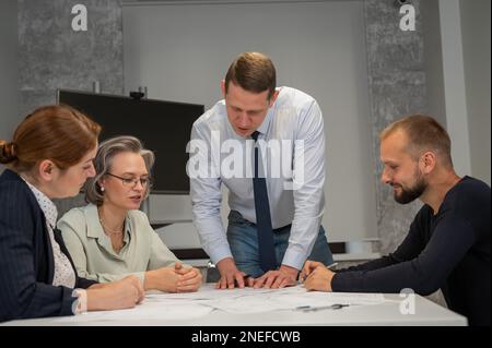 Ein weißer Mann steht und ändert eine Zeichnung, drei Kollegen sitzen an einem Tisch und hören ihm zu. Stockfoto