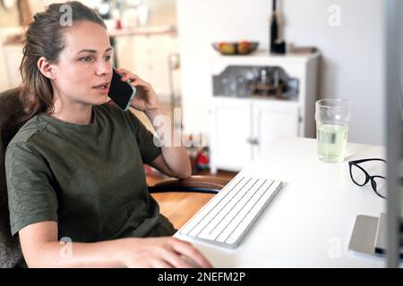 Junge Frau, die von zu Hause aus arbeitet, am Computer arbeitet und mit dem Handy telefoniert. Stockfoto