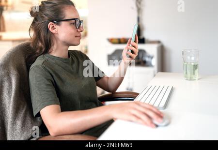 Frau mit Brille, die von zu Hause aus arbeitet, sitzt am Schreibtisch und benutzt ihren Computer und ihr Mobiltelefon. Stockfoto
