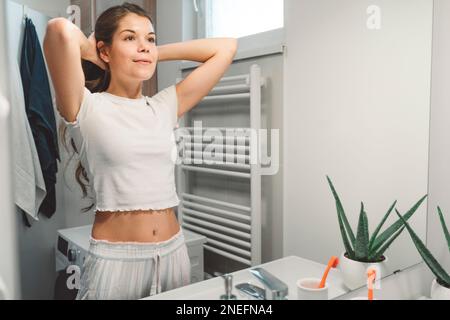Junge Weiße, die sich die Haare im Pferdeschwanz bindet, während sie sich im Spiegel ansieht Stockfoto