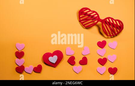 Das Konzept des Valentinstages. Die Inschrift Liebe ist auf einem gelben Hintergrund mit kleinen rosa und roten Herzen angelegt. Blick von oben. Rote Brillen schon Stockfoto