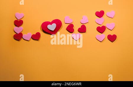 Das Konzept des Valentinstages. Die Inschrift Liebe ist auf einem gelben Hintergrund mit kleinen rosa und roten Herzen angelegt. Draufsicht. Mit Kopierbereich. Stockfoto