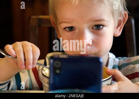 Ein kleiner Junge, der während des Mittags oder Abendessens am Tisch das Telefon benutzt. Kinder essen Essen und sehen sich Cartoon auf dem Smartphone an, weil Nomophobie oder Stockfoto