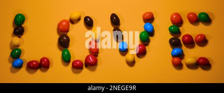 Konzept des Valentinstages. Flache Grußkarte. Bunte süße Süßigkeiten mit Buchstaben und Love-Inschrift auf gelbem Hintergrund. Stockfoto