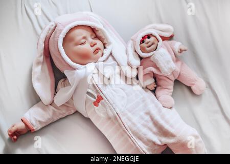 Ein kleines Baby in einem Osterhase-Kostüm schläft auf einem weißen Bett und verteilt Jahre an den Seiten neben einer hausgemachten Puppe in einem Overall mit Kaninchenohr. Stockfoto
