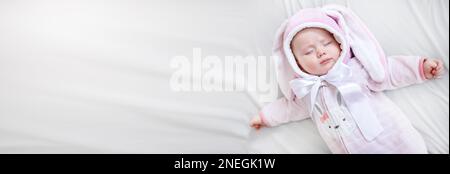 Das Baby schlief auf einem paska in Overalls im Schlafzimmer auf einem weißen Bett ein. Das kleine Mädchen schläft ruhig. Ruhiger tiefer, ruhiger Schlaf des Kindes Stockfoto