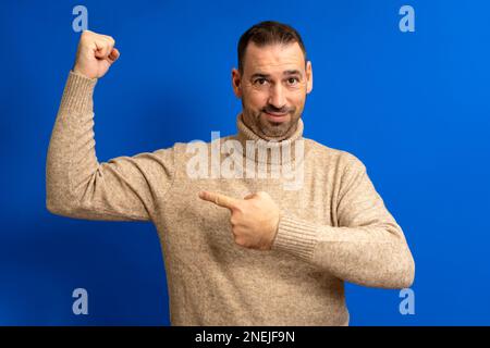 Porträt lateinischer bärtiger Mann, der mit dem Finger auf seinen Armmuskel zeigt, Rabatt, der mit zufriedenem Gesicht auf die Kamera schaut. Innenstudio, isoliert auf Blau Stockfoto