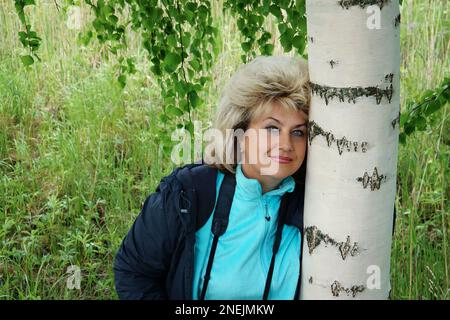 55-60-jährige Frau steht vor dem Hintergrund von Birkenästen. Großes Porträt einer russischen Frau mit blondem Haar. Stockfoto