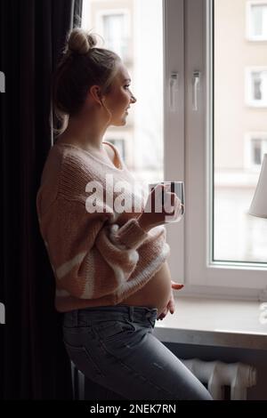 Charmante, junge, schwangere Frau in pinkfarbenem Strickpullover blickt mit einem Lächeln aus dem Fenster. Fürsorge und Mutterschaft. Stockfoto