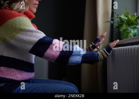 Frau Zu Hause In Handschuhen Wärmende Hände Durch Heizkörper Während Der Lebenshaltungskosten-Energiekrise Stockfoto