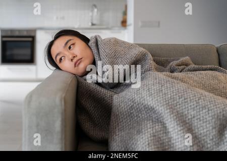 Frustrierte asiatische Frau, die auf der Couch liegt, sieht leider durch Depressionen gewickelt in Karo zu Hause aus. Stockfoto