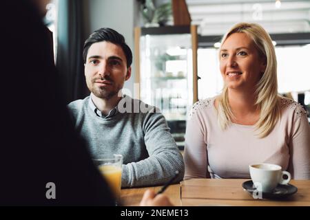 Lächelndes weißes Paar, das am Schreibtisch gegenüber seinem Finanzberater sitzt und genau zuhört, was er sagt Stockfoto