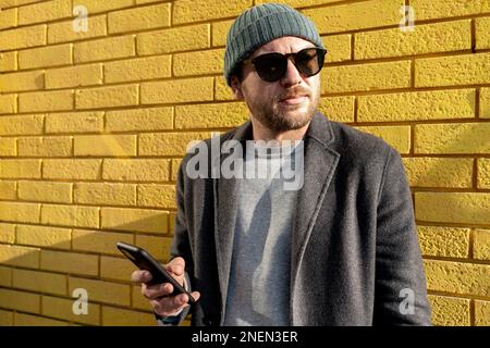 Ein Mann mit Hut und Sonnenbrille steht an einer gelben Ziegelwand und hält ein Handy in der Hand. Stockfoto