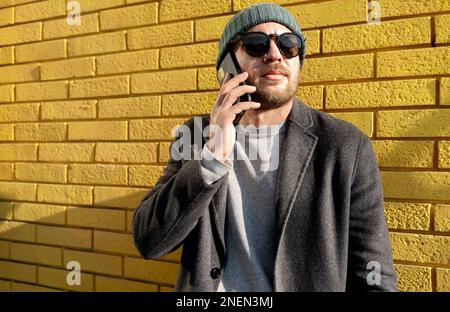 Urbaner Lebensstil Erwachsener mit Hut und Sonnenbrille, der an einer gelben Backsteinwand steht und am Handy anruft. Stockfoto