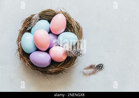Happy Easter Konzept. Vorbereitung auf den Urlaub. Farbenfrohe, pastellfarbene Ostereier im Nest mit Federn auf betonsteingrauem Hintergrund. Einfacher Minimalismus flach Lay Draufsicht Kopieplatz Stockfoto