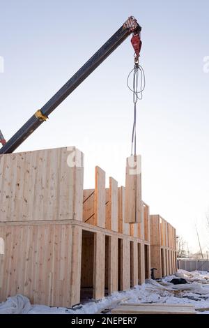 Der Bau eines neuen modernen modularen Hauses aus Verbundplatten an einem sonnigen Wintertag. Ein Kran hebt eines der Bleche an Stockfoto