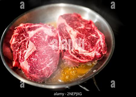 Edelstahlpfanne und Nahaufnahme von zwei rohen Ribeye-Fleischsteaks, die auf dem Herd in Ölfett und schwarzem Hintergrund mit fettartiger Marmorierung gekocht werden Stockfoto