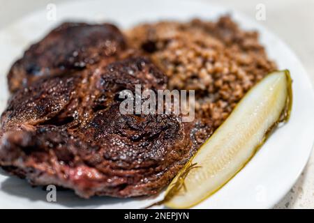 Gekochtes Ribeye Steak Makro-Nahaufnahme mit dickem Schnitt und brauner Kruste auf weißem Teller mit Dill-Gurke und Buchweizenkörn im Hintergrund Stockfoto